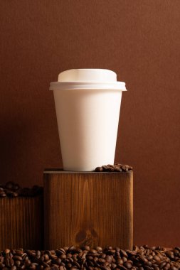 Kahverengi arka planda, kahve çekirdekleri ile plastik kapaklı, tek kullanımlık boş kağıt bardak.
