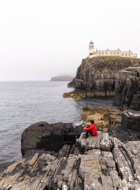Photo of a woman wearing a red coat on top of the rocky cliff near the Neist Point lighthouse in the Highlands, Scotland during a summer day.
