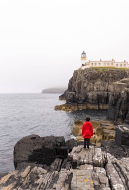 Photo of a woman wearing a red coat on top of the rocky cliff near the Neist Point lighthouse in the Highlands, Scotland during a summer day.