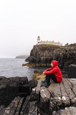 Portrait of a woman wearing a red coat and sitting on top of the rocky cliff near the Neist Point lighthouse in the Highlands, Scotland during a summer day.