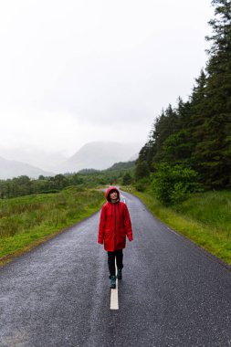 Portrait of a woman wearing a red raincoat while walking alone on a road that crosses a valley surrounded by mountains and fir trees during a foggy day in the highlands, Scotland