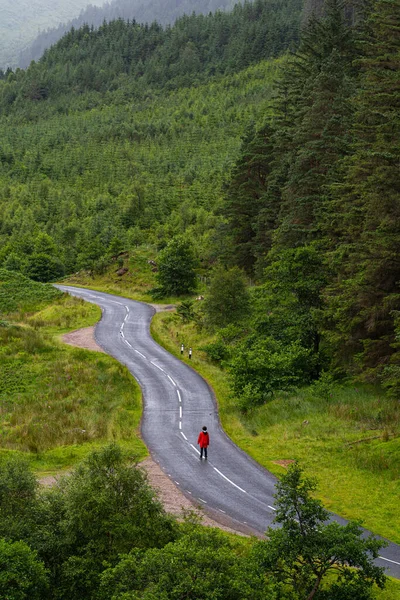 Photo of a person walking alone on a road that crosses a valley surrounded by mountains and fir trees during a foggy day in the highlands, Scotland