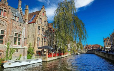 Brugge, Belçika - 9 Ekim. 2021: Köprüdeki su kanalına (Nepomucenusbrug) yeşil ağaç, restoran ve ortaçağ evleriyle açık mavi sonbahar gökyüzüne karşı bakın