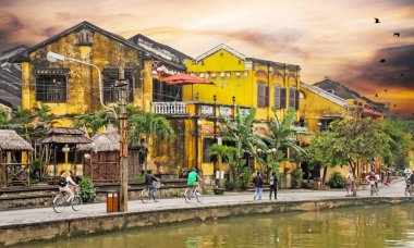 Hoi An, Vietnam - 9 Aralık. 2015: Eski sarı tipik evler, bisikletçilerle nehir geçidi, yağmur mevsiminde bulutlu mistik günbatımı gökyüzü