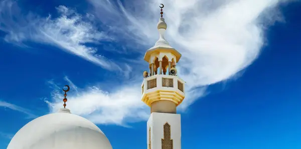Symbols of islam and muezzin prayer call concept: isolated white dome, minarette tower with loudspeakers, islamic crescent moon finial symbol against blue sky, Oman
