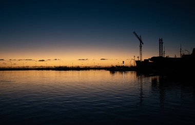 Güneş denizin üzerinde ufuktan batıyor. Vinçli bir sanayi limanının gölgeleri ön planda görülebilir. Deniz sakin ve karanlık..