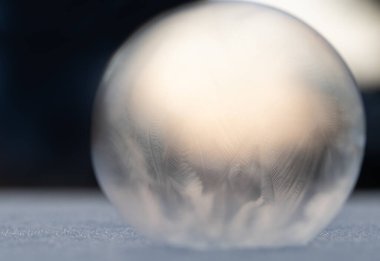 Donmuş sabun köpüğünün yakın çekimi. Top karın üzerinde yatıyor. Arkaplan karanlıktır ve arada parlak bir ışık vardır. Kabarcık şeffaf