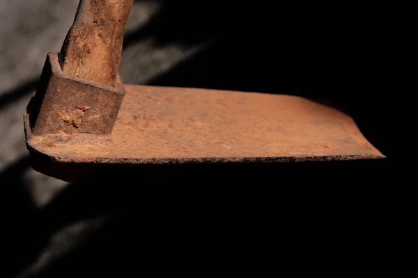 Close up of a rusty spade hovering above the ground from above. The background is light and shadow. The handle is made of wood.