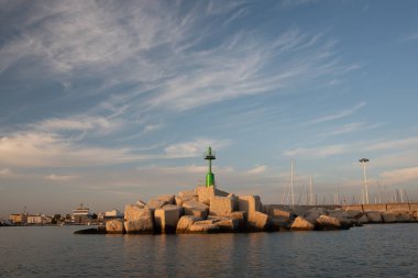 At the port of Mazara del Vallo in Sicily, a small green lighthouse stands on concrete breakwaters in the evening sun. The sky is blue with clouds. The tower is reflected in the water.
