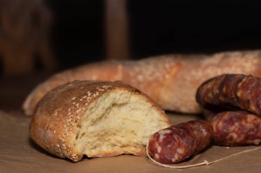 Masanın üzerinde susam tohumlu beyaz ekmek var. Ekmek dilimlendi. Önünde de kurutulmuş salam var, o da kesiliyor. Arka planda tahtadan bir sandalyenin silüeti var..