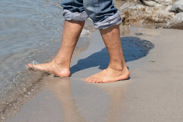 白い男の素足が浜辺の水の方へ歩いていく その男はジーンズをはいてしまった テキストのスペースがあります ストックフォト