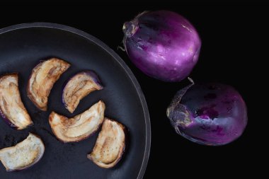 Dökme demir bir tavada duran kızarmış patlıcanların yakın çekimi. Taze mor patlıcanlar tavanın yanında. Arka plan karanlık.