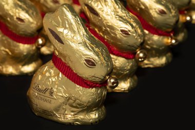 Bir grup Lindt çikolatalı tavşanı karanlık bir arka plana karşı duruyor. Tavşanlar yan yana ve arkalarında dizilmişler. Yan taraftan çekiliyorlar..