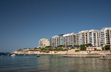 Malta sahilinde yeni inşa edilmiş gökdelenler denize yansıyor. Gökyüzü mavi. Güneş parlıyor. İnsanlar binaların önünde güneşleniyorlar..
