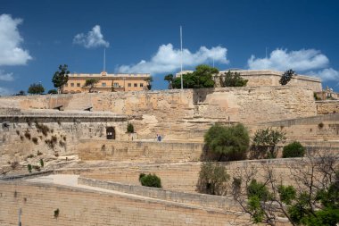 Turistler Valletta şehir duvarlarından aşağı doğru yürüyorlar. İniş, yüksek kireçtaşı duvarın üzerinden yılanlarla yapılıyor. Gökyüzü beyaz bulutlarla mavidir