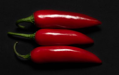 Üç taze kırmızı kapsül koyu bir arka planda yan yana uzanıyor. Kırmızı biberler ışıkta parlıyor.
