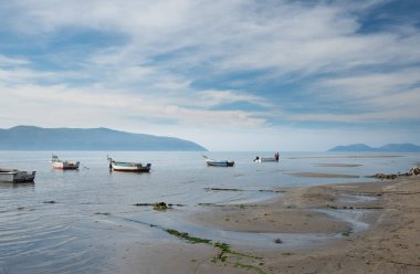 Küçük tekneler denizin ortasındaki plaja demirlediler. Gökyüzü bulutlu. Ön planda kum var. Arka planda küçük adaların bulanık hatları.