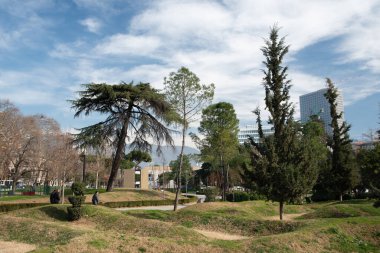 Arnavutluk 'un Tiran kentindeki parktan insanlar geçiyor. Çayır tepeliktir. Arada ağaçlar var. Arka planda, gökdelenler ve bulutlu mavi gökyüzü.