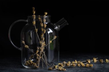 Bir cam sürahi koyu bir arka planın önünde duruyor. Sürahide bir sürü albanya çayı var. Tencerenin yanında da çayın çiçekleriyle kurumuş dallar var..