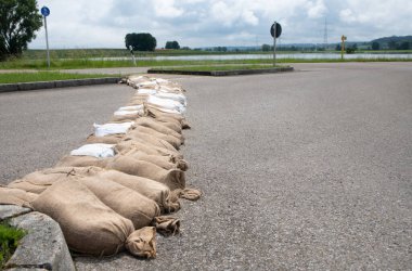 Kum torbaları, sel tehdidine karşı erişim yolunu korumak için bir yola yerleştirildi. Arka planda sel basmış bir çayır ve gökyüzü bulutlarla dolu..