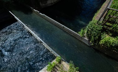 Dar bir nehrin yukarıdan başka bir kanal üzerindeki köprünün üzerinden geçişini izle. Suyun yanında yeşil bitkiler yetişir..