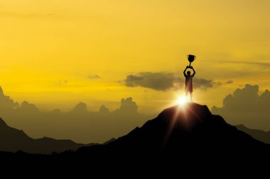 Siluet işadamı dağın tepesinde duruyor ve iş dünyasının kazananı için güneş ışığı altında bir kupa tutuyor. Başarılı ve hedeflenen hedef kavramı elde ediyor..
