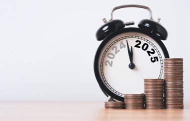 2024 ve 2025 yıllarında ahşap masa üzerinde siyah alarm saati ve yeni yıl konseptinin mali hazırlıkları için birikmiş madeni paralar.