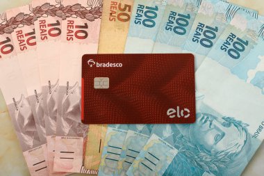 Bradesco card with elo logo, red card with chip on top of Brazilian money clipart