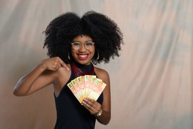 Genç Afrikalı Amerikalı kadın bej arka planda Brezilya parası tutuyor.,