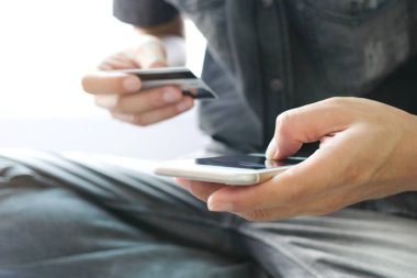 Genç adam oturma odasında kanepede oturdu, telefonunu, akıllı telefonunu ve kredi kartını internetten satın almak için kullandı.