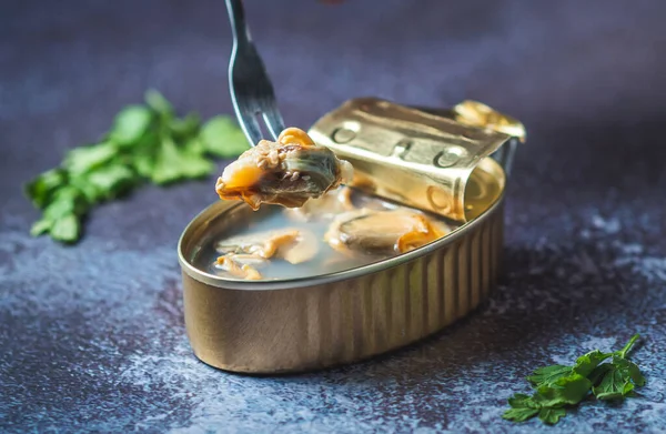 Fork pricking canned clams on a dark blue table with parsley. Ready for eat. 
