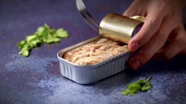 Hand with a fork picking up canned mackerel on a dark blue table with parsley. Ready to eat.