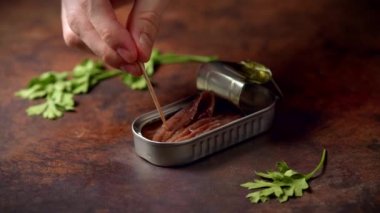 Hand with a toothpick picking up canned anchovies on a dark blue table with parsley. Ready to eat.