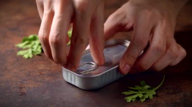 Hands opening a can of sardines on a dark brown table with parsley. Ready to eat.