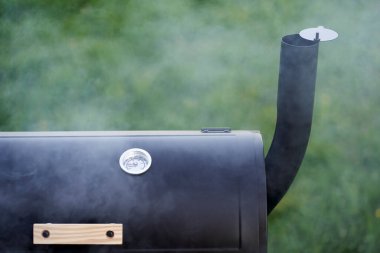 Close up of new smoker grill surrounded by steam. In the green nature.