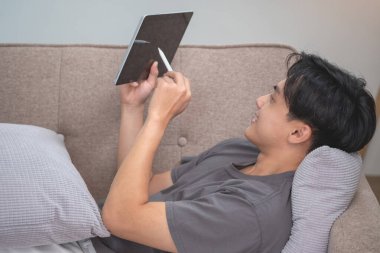 Elinde tabletle müzik dinleyen beyaz bir adamın portresi. Evde tatilde dinlenmek için evin içindeki kanepede bir kalem tutuyor..