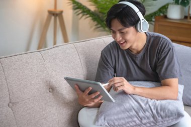 Elinde tabletle müzik dinleyen beyaz bir adamın portresi. Evde tatilde dinlenmek için evin içindeki kanepede bir kalem tutuyor..