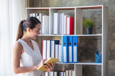 Asyalı iş kadını iş verilerini grafiklerle, sarı defterle el ele tutuşan grafiklerle ve ofisteki masada duran iş belgeleriyle sunuyor..