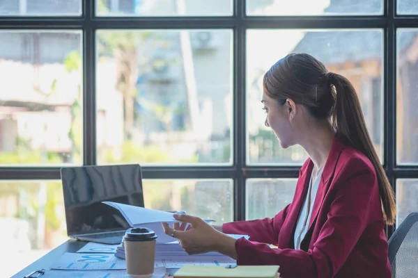 stock image Financial, Asian businesswoman in red suit holding cup of coffee sitting on desk in office, having computer for doing accounting work at workplace to calculate annual profit by duty, Business idea