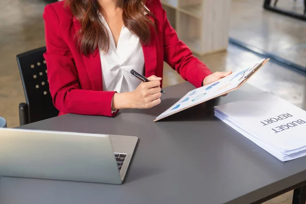Asian businesswoman in red suit holding wooden folder analyzing accounting work on laptop computer on the desk in office.
