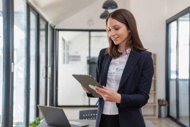 Siyah takım elbiseli şık bir beyaz iş kadını elinde cep telefonu, tablet ve dizüstü bilgisayarla müşterilerin iletişime geçmesini bekliyor..