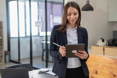 Siyah takım elbiseli şık bir beyaz iş kadını elinde cep telefonu, tablet ve dizüstü bilgisayarla müşterilerin iletişime geçmesini bekliyor..