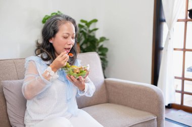 Asyalı son sınıf öğrencisi, sağlıklı sebze salatası yiyor ve sağlıklı bir vücut için evdeki kanepede mutlu bir şekilde yemek yiyor. sağlıklı gıda konsepti