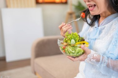 Asyalı son sınıf öğrencisi, sağlıklı sebze salatası yiyor ve sağlıklı bir vücut için evdeki kanepede mutlu bir şekilde yemek yiyor. sağlıklı gıda konsepti