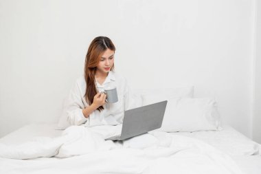 Beyaz gecelikli bir kadın hafta sonu uyanıp dinleniyor ve cep telefonuyla oynuyor ekmek yiyor ve beyaz yatak odasında çay içiyor. Sabah tatili.