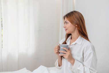 Beyaz gecelikli bir kadın hafta sonu uyanıp dinleniyor ve cep telefonuyla oynuyor ekmek yiyor ve beyaz yatak odasında çay içiyor. Sabah tatili.