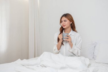 Beyaz gecelikli bir kadın hafta sonu uyanıp dinleniyor ve cep telefonuyla oynuyor ekmek yiyor ve beyaz yatak odasında çay içiyor. Sabah tatili.