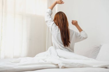 Beyaz gecelikli bir kadın hafta sonu uyanıp dinleniyor ve cep telefonuyla oynuyor ekmek yiyor ve beyaz yatak odasında çay içiyor. Sabah tatili.