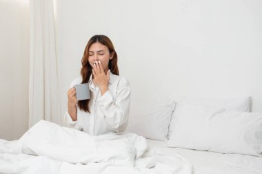 Beyaz gecelikli bir kadın hafta sonu uyanıp dinleniyor ve cep telefonuyla oynuyor ekmek yiyor ve beyaz yatak odasında çay içiyor. Sabah tatili.