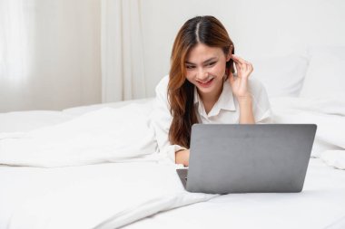 Beyaz gecelikli bir kadın hafta sonu uyanıp dinleniyor ve cep telefonuyla oynuyor ekmek yiyor ve beyaz yatak odasında çay içiyor. Sabah tatili.